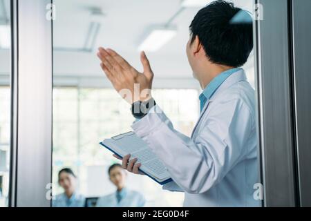 Rückansicht der jungen männlichen Arzt hält Zwischenablage zeigt in Richtung Glastür bei einem Treffen mit Kollegen im Krankenhaus Konferenzraum Stockfoto