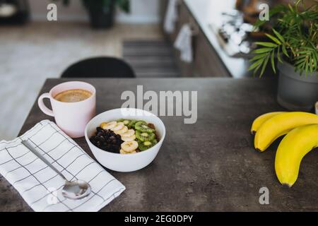 Rosa Kaffeetasse, Schüssel mit gehackten tropischen Früchten Kiwi und Banane, Heidelbeeren, Löffel auf der Theke in der stilvollen Loft-Küche. Unscharfer Hintergrund. Hig Stockfoto