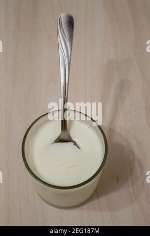 Ein zylindrischer Glasbehälter mit frisch zubereitetem hausgemachtem Joghurt mit gefrorenem Schaum um den Rand mit einem Metallteelöffel in seiner Oberfläche installiert, c Stockfoto