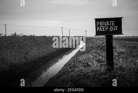 Ein privates Keep-Out-Schild in einem landwirtschaftlichen Feld in den Lincolnshire Fens neben einem Drainagesteich. Düster und schwarz-weiß Stockfoto