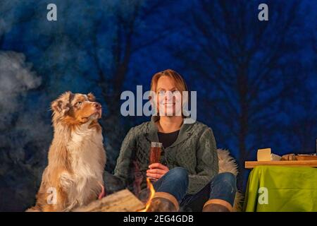 Junge Frau, die am Lagerfeuer sitzt und Wein trinkt. Ihr australischer Schäferhund sitzt neben ihr und genießt sich. Draußen im Park bei Blau Stockfoto