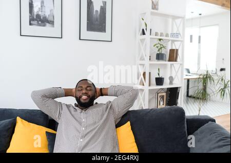 Zufriedene junge afrikanische Mann ruht auf bequeme Couch in weißen gemütlichen modernen Wohnzimmer, die Hände hinter dem Kopf, Millennial Hipster Kerl genießen keine Stockfoto