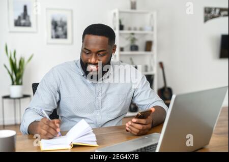 Lächelnder afrikanischer Freiberufler, der Notizen macht, mit dem Laptop arbeitet, das Notebook vor dem Laptop anschaut, am Telefon chattet Stockfoto