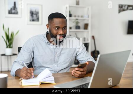 Fokussierte glückliche junge afroamerikanische Mann Student Freiberufler Notizen machen Studium mit Laptop arbeiten, tragen airpods Blick auf den Bildschirm des Stockfoto
