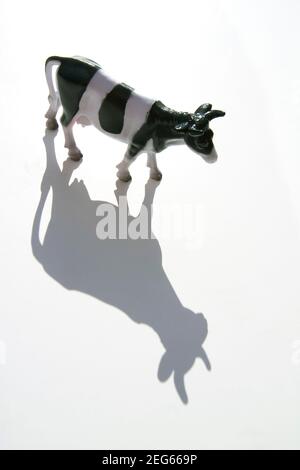 Schwarz-weiße Spielzeugkuh, die bei hellem Licht einen verspielten Schatten auf eine weiße Oberfläche wirft Stockfoto