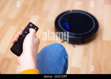 Mann ruht sich aus, während der Roboter-Staubsauger Aufgaben erledigt, zu Hause sauber arbeitet. Man steuert einen Roboter-Staubsauger mit einer Fernbedienung. Reinigungsroboter V Stockfoto