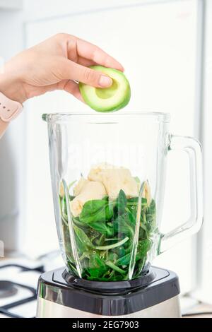 Zutaten für grüne Detox-Smoothies im Mixer. Spinat, Banane und Avocado. Seitenansicht. Hand füllt einen Mixer oder Mixer mit gesunden Zutaten: av Stockfoto