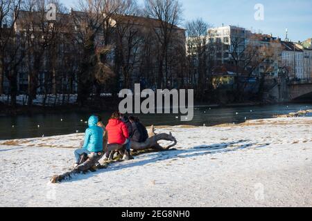 München, Deutschland - 13. Februar 2021: Menschen, die auf dem Baumstamm an der Isarseite sitzen, genießen einen sonnigen Tag im Freien Stockfoto