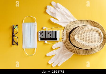 Reise nach Covid-19 und neue normale Konzepte. Draufsicht auf medizinische Gesichtsmaske, Handschuhe, Kopfhörer mit Telefon und Strandhut auf gelbem Hintergrund schließen U Stockfoto