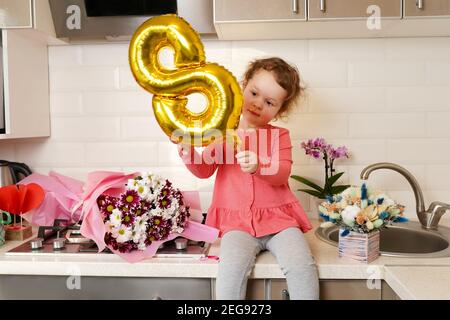 Nette lustige kleine Mädchen hält goldenen Ballon 8 acht sitzen auf Küchentisch mit Blumensträußen des Frühlings Blumen, Gruß Mutter oder Großmutter zu Hause Stockfoto