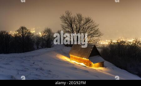Fantastische Winterlandschaft mit glühendem Holzhaus vor dem Hintergrund glühender Lichter der Stadt im Nebel. Gemütliche Hütte in den Karpaten. Weihnachtsfeiertagskonzept Stockfoto