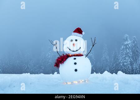 Lustiger Schneemann in stylischem roten Hut und rotem Skalf auf verschneiten Bergen. Leuchtende Girlande für Weihnachtsstimmung Stockfoto