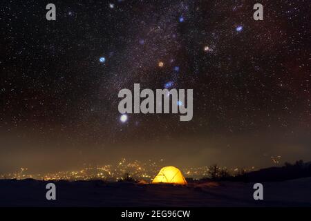 Gelbes Zelt, beleuchtet von innen vor dem Hintergrund der leuchtenden Lichter der Stadt und des unglaublichen Sternenhimmels. Tolle Nachtlandschaft. Touristen campen in den Winterbergen. Reisekonzept Stockfoto