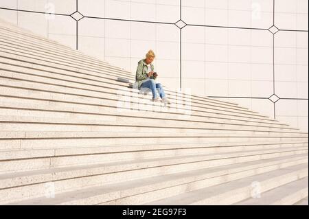 Minimalismus in der Architektur. Ein junges Mädchen in Jeans und ein grüner Windbreaker mit blondem Haar sitzt auf weißen Steintreppen und schaut Fotos in der Kamera an. Stockfoto