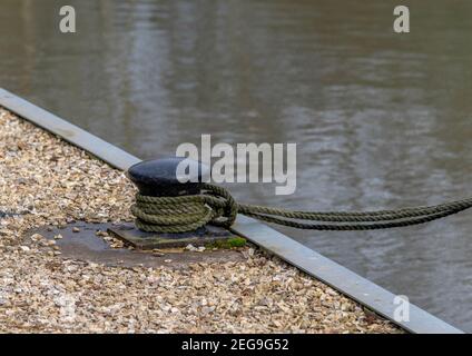 Seil gebunden an einem schwarzen Ankerplatz Poller auf dem Dock mit Wasser im Hintergrund. Stockfoto