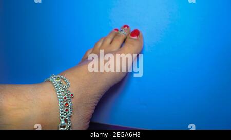 Die traditionelle hinduistische Braut trägt Paaje (Anklets) in Indien. Weiblicher Fuß mit Nagellack isoliert auf blauem Hintergrund Stockfoto
