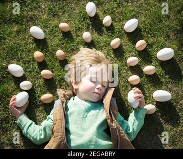 Viele weiße und beige Hühner- und Gänseeier liegen um den Kopf eines 4-jährigen blonden Jungen, der auf dem Gras schläft. Fröhliche Vorbereitung auf EAS Stockfoto