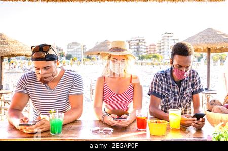 Multirassische Freunde mit geschlossenen Gesichtsmasken mit Tracking-App mit Mobile Smartphones - gelangweilte junge Menschen am Strand Cocktail Bar Stockfoto