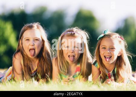 Drei kleine Mädchen, die ihre Zungen herausstreckt und im Park auf dem Gras liegen. Kleine Rebellen. Schwesternschaft. Stockfoto