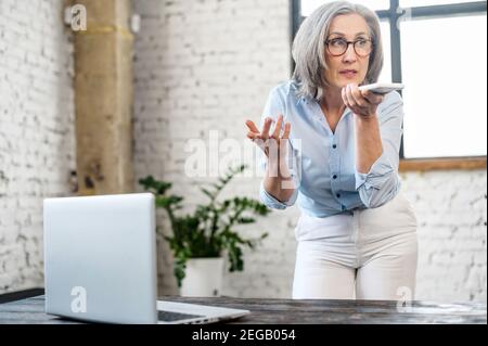 Beschäftigte ältere Geschäftsfrau in Brillen mit Spracherkennungs-App auf dem Telefon. Moderne gealterte weibliche Büroangestellte hält Smartphone in der Nähe von Mund und Stockfoto