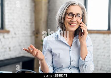Lächelnde und positive Geschäftsfrau, die im modernen Büro mit dem Smartphone spricht. Eine ältere Verkäuferin telefoniert. Reife Hündin Stockfoto