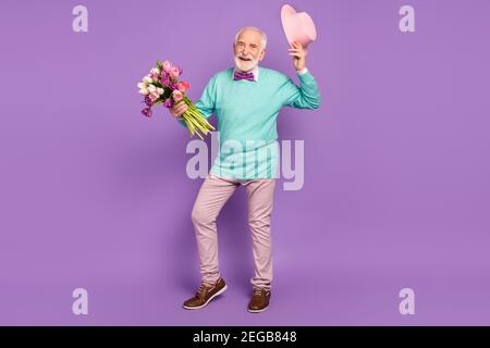 Foto in voller Größe von positiven Mann halten Blumen Hand heben Kopfbedeckung strahlendes Lächeln isoliert auf violettem Hintergrund Stockfoto