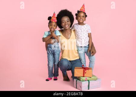 Portrait von jungen glücklich afrikanischen Mama, Umarmung ihrer beiden niedlichen kleinen Kinder Töchter in Party-Hüte, Geburtstagsfeier auf pastellrosa feiern Stockfoto