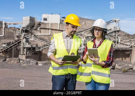 Seriöse professionelle Vorarbeiter mit Clipboard diskutieren Projekt mit weiblichen Ingenieur Mit Tablet während der Zusammenarbeit auf Industriebau sind Stockfoto