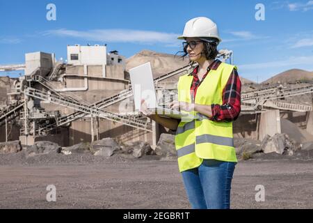 Seriöse Ingenieurin in Hardhat und Weste suchen Informationen über Laptop, während er in der Nähe der Industrieanlage der Baustelle steht Stockfoto