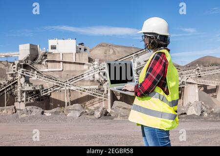 Seitenansicht der Ingenieurin bei der Arbeit mit Hut und Weste Auf Laptop mit schwarzem Bildschirm beim Besuch Baustelle von Unvollendete industrielle fa Stockfoto