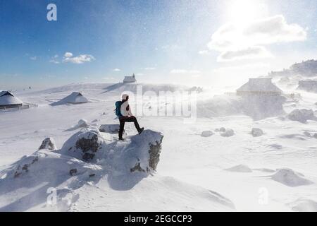 Frau, die bei windigen Bedingungen zur Kapelle der Schneemarie auf der Velika planina-Wiese in Slowenien wandert Stockfoto