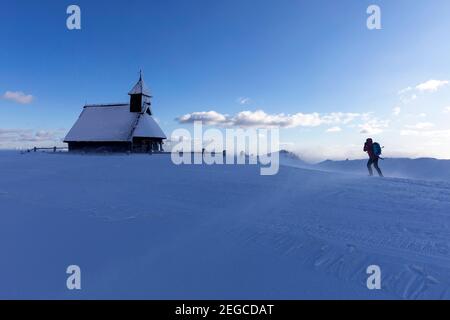 Frau, die bei windigen Bedingungen zur Kapelle der Schneemarie auf der Velika planina-Wiese in Slowenien wandert Stockfoto