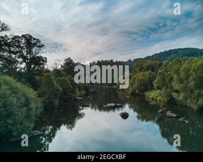 Bunte Landschaft mit kleinem Fluss voller großer Steine dazwischen Büsche und Bäume am bewölkten Morgen Stockfoto