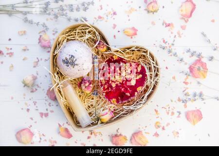 Flache Ansicht von verschiedenen hausgemachten Spa-Geschenken: Rosa Seife mit Rosenblüten, Badebombe mit Lavendel und Lippenbalsam in Pappschachtel. Stockfoto