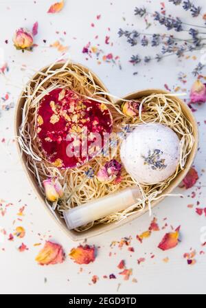 Flache Ansicht von verschiedenen hausgemachten Spa-Geschenken: Rosa Seife mit Rosenblüten, Badebombe mit Lavendel und Lippenbalsam in Pappschachtel. Stockfoto