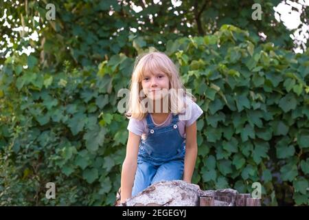 Ein blondes Mädchen sitzt auf einem Stein vor einem Hintergrund von dichten grünen Bäumen. Sommerurlaub mit meiner Großmutter im Dorf. Stockfoto