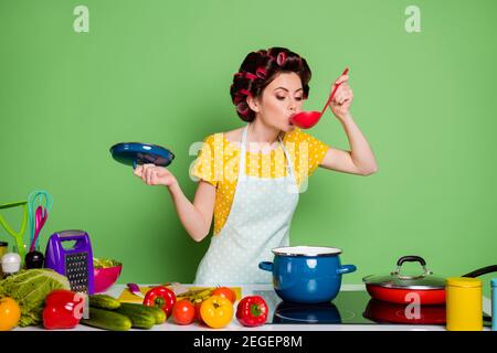 Foto Retro stilvolle Mädchen Koch bereiten vegetarische Suppe kochen Sauce Pfanne versuchen essen Schöpflöffel Tisch Tomate Gurke tragen Haarrollen Gepunkteter gelber Rock Stockfoto