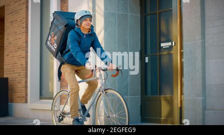 Handsome Happy Food Delivery Mann trägt Thermal Rucksack reitet sein Fahrrad durch die stilvolle moderne City District. Schuss lächelnder Courier Stockfoto