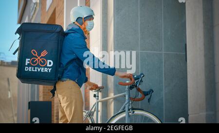 Lebensmittel-Lieferung Mann trägt schützende medizinische Gesichtsmaske und Thermal-Rucksack auf einem Fahrrad liefert Bestellung an einen Kunden. Kurier Liefert Takeaway Lunch Stockfoto