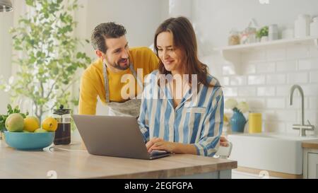 Schöne junge Paar mit Laptop-Computer in der Küche zu Hause. Schlafanzug Für Frauen. Mann trägt eine Schürze ist ermutigend Freundin mit einem Stockfoto