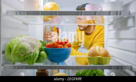 Innerhalb der Küche Kühlschrank: Junge Bartmann nimmt aus Wasserflasche. Gesunder Mann Bleibt Hydratisiert. Point of View POV Aufnahme aus dem Kühlschrank voller Stockfoto