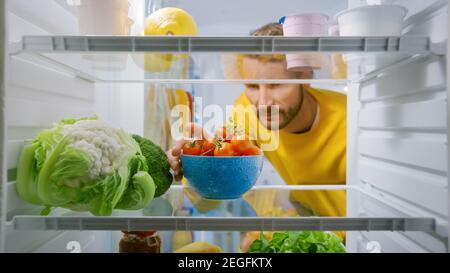 Innenküche Kühlschrank: Handsome man nimmt Kirschtomaten aus dem geöffneten Kühlschrank. Mann, Der Eine Gesunde Mahlzeit Zubereitet. Blickwinkel POV Aufnahme aus dem Kühlschrank Stockfoto