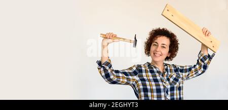 Das junge kaukasische Mädchen mit lockigem Haar hält ein Holzbrett und einen Hammer. Sie hebt ihre Arme, um ihm zu zeigen, wie man zelte. Das Innere einer Wohnung. Sie weiße Wand. Stockfoto