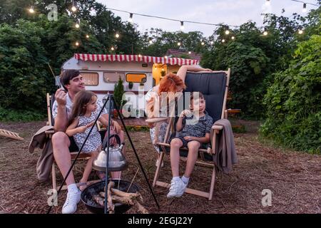 Fröhliche Familie auf Picknick außerhalb der Stadt, Würste auf Spieße werden über Feuer gebraten. Sitzen auf hölzernen Liegestühlen an der Anhängerbahn. Eltern sind ein Narr Stockfoto