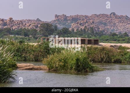 Anegundi, Karnataka, Indien - 9. November 2013: Grauer Tungabhadra-Fluss. Gebäude Ruinen neben Sooryanarayana Tempel Komplex auf seiner grünen Insel un Stockfoto