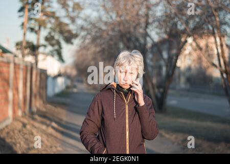 Eine schöne Frau mittleren Alters in legerer Kleidung spricht am Telefon mit einem Lächeln auf ihrem Gesicht. Abends in der Sonne durch die Stadt spazieren. Stockfoto
