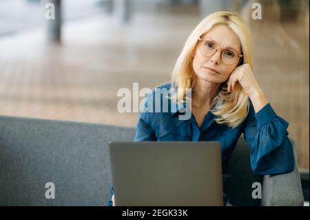 Serious reife blonde-Haar Geschäftsfrau sitzt auf dem Sofa im Büro. Elegante einflussreiche Dame schaut direkt auf die Kamera, fühlt sich müde, überarbeitetes Konzept Stockfoto