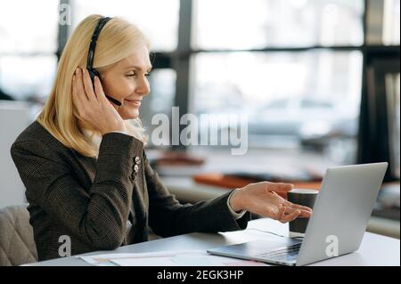 Kaukasische Managerin im Headset schaut auf den Laptop-Bildschirm und führt Online-Geschäftstreffen durch. Reifer Mitarbeiter, der mit Kollegen über Arbeitsfragen spricht, Kommunikationskonzept Stockfoto