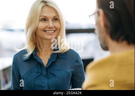 Attraktive blonde-Haare Geschäftsfrau, die ein Gespräch mit männlichen Kollegen, lächelnd. Mitarbeiter mittleren Alters auf einer Pause im modernen Büro, Gespräch, Kommunikationskonzept Stockfoto