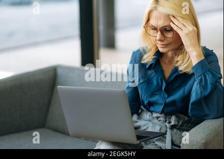 Reife elegante Frau in Brillen sitzen auf dem Sofa und haben Kopfschmerzen. Erschöpft kaukasischen Geschäftsfrau ist mit Laptop, Gefühl müde nach Brainstorming, Überarbeit Konzept Stockfoto